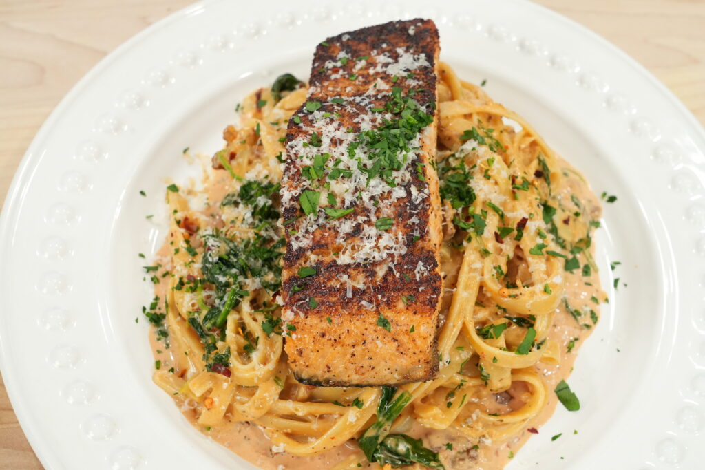 Photo of completed and plated Marry Me Salmon Dish by Mr. Make It Happen. The fettuccine pasta is plated and garnished with parsley with a large piece of salmon placed on top garnished with parmesan cheese and parsley.