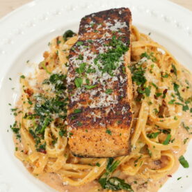 Photo of completed and plated Marry Me Salmon Dish by Mr. Make It Happen. The fettuccine pasta is plated and garnished with parsley with a large piece of salmon placed on top garnished with parmesan cheese and parsley.