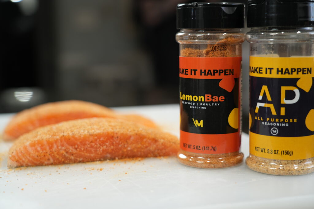 2 pieces of raw cut salmon on a cutting board with 1 bottle of Mr. Make It Happen's Lemon Bae Seasoning and 1 bottle of Mr. Make It Happen's All-Purpose Seasoning