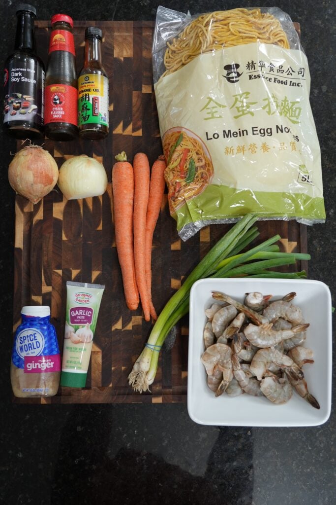 Raw shrimp, fresh egg noodles, carrots, onion and green onions laid out on a wood board